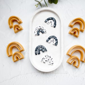 May include: A set of six rainbow-shaped cookie cutters. Five of the cutters are black and white with a floral pattern. One cutter is a solid yellow.