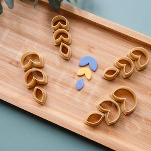 Puede incluir: Un juego de seis cortadores de galletas con forma de hojas y flores. Los cortadores están hechos de plástico marrón claro y están dispuestos sobre una superficie de madera.