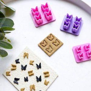 May include: Four sets of butterfly-shaped cookie cutters, two pink, one purple, and one brown. The cutters are arranged on a white surface with a sprig of green leaves in the upper left corner. The brown cutter has been used to make several small butterfly shapes on a beige surface.