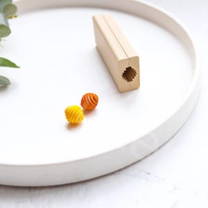 May include: A wooden mold for making small, spiral-shaped pasta. Two pieces of pasta, one orange and one yellow, are on a white surface next to the mold.