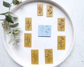 May include: A collection of floral-themed stamps and a blue mold are arranged on a white surface. The stamps are rectangular, with detailed botanical designs in a light brown color. The blue mold features similar floral patterns. Green leaves are in the background.