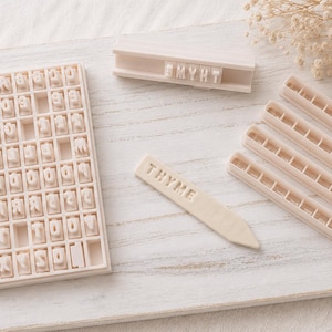 May include: A set of off-white plastic tools for stamping text, including a rectangular grid tray filled with individual letter stamps, a long holder with the letters THYME, and four pointed stakes, one of which is embossed with the word THYME.
