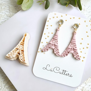 以下が含まれることがあります： エッフェル塔型の薄いピンク色のイヤリングと、お揃いのクッキーカッター。イヤリングは、クリアクリスタルが付いたゴールドカラーのポストです。クッキーカッターは薄いベージュ色です。「Lü Cutters」と書かれたカード。