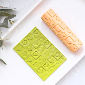 Puede incluir: Dos rodillos para hornear, uno verde y uno naranja, con un patrón de corazón.
