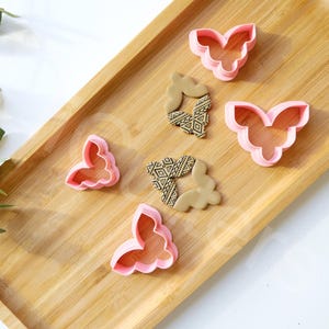 Puede incluir: Una bandeja de madera con cuatro cortadores de galletas en forma de mariposa rosa y dos cortadores de galletas con patrones geométricos dorados.
