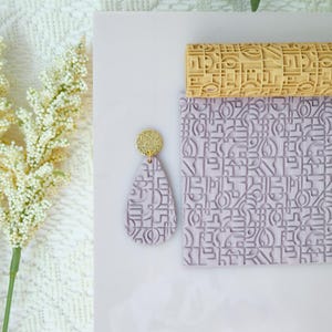May include: A light purple clay earring with a gold glitter top, next to a matching clay roller and sheet. The earring and sheet have a geometric pattern. A yellow clay roller with a similar pattern is also present.