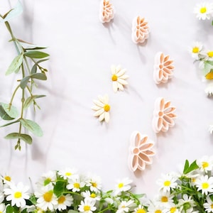 Pode incluir: Um conjunto de seis cortadores de biscoitos de plástico branco em forma de flor sobre um fundo branco com margaridas brancas espalhadas ao redor.