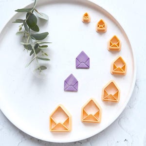 May include: A set of nine envelope-shaped cookie cutters in various sizes. The cutters are arranged in a circle on a white plate. The cutters are a light orange color, except for two that are purple.