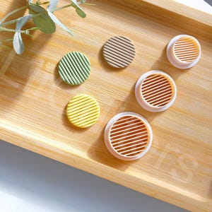 May include: Five round cookie cutters on a wooden surface. The cutters are in different colours and patterns, including green, yellow, brown, and pink.