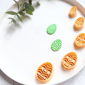 Pode incluir: Um prato branco com seis cortadores de biscoitos de ovo de Páscoa de plástico laranja e verde. Os cortadores têm um padrão ondulado.