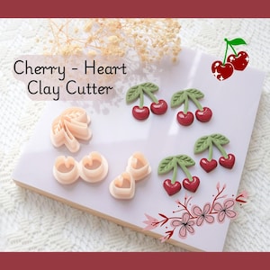 Op de afbeelding: Een verzameling kleisnijders en kleikersen op een wit oppervlak. De snijders hebben de vorm van kersen en harten. De kersen zijn rood met groene bladeren. De tekst "Cherry - Heart Clay Cutter" is zichtbaar.