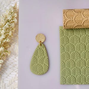 May include: A single green teardrop-shaped earring with a gold-colored circular top. The earring has a leaf pattern. Next to the earring are two textured clay stamps, one rectangular and one cylindrical, with a similar leaf pattern.