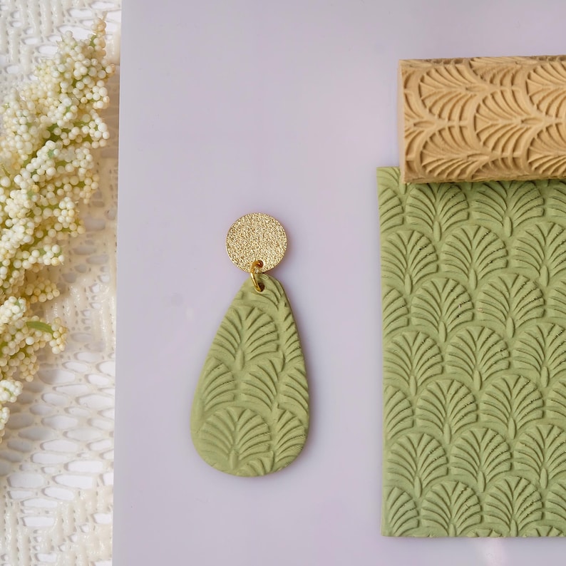 May include: A single green teardrop-shaped earring with a gold-colored circular top. The earring has a leaf pattern. Next to the earring are two textured clay stamps, one rectangular and one cylindrical, with a similar leaf pattern.
