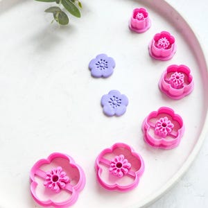 Puede incluir: Una colección de cortadores de galletas y moldes de fondant con forma de flor rosa y morada. Los cortadores son de varios tamaños y presentan un diseño floral detallado. Los artículos están dispuestos sobre una superficie blanca, lo que sugiere un tema de repostería o manualidades.