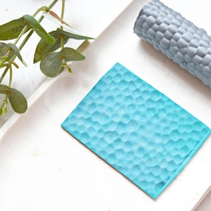 May include: Two silicone molds for creating textured surfaces. One mold is a flat rectangle with a blue textured surface. The other mold is a cylinder with a gray textured surface.