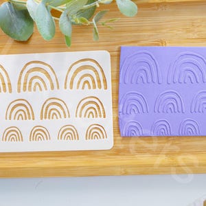 Puede incluir: Una plantilla de plástico blanco con un patrón repetitivo de arcos de arcoíris. La plantilla está junto a una lámina de silicona morada con el mismo patrón de arco de arcoíris en relieve.