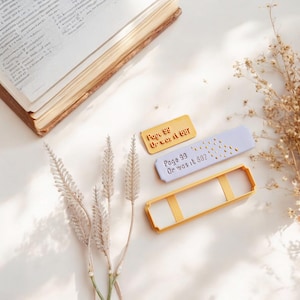 Könnte beinhalten: Ein lila und gelber Stempel aus Kunststoff mit dem Text "Seite 99 Oder war es 68?" und ein zweiter Stempel mit dem Text "Seite 99 Oder war es 60?" mit goldenen Glitzerakzenten.