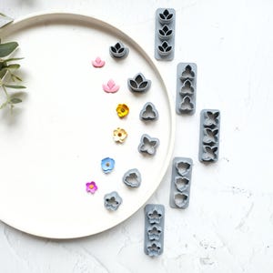 Könnte beinhalten: Ein Set aus grauen Kunststoff-Ausstechern in Form von Blumen und Blättern auf einem weißen Teller mit verschiedenen bunten, blumenförmigen Fondant-Dekorationen.