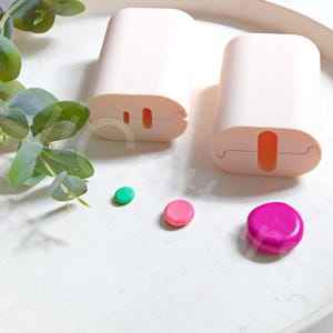 May include: Two light pink, rectangular cases with rounded edges and a slot. Three colorful, round objects in green, pink, and magenta are scattered nearby. A sprig of green leaves adds a natural touch to the composition.