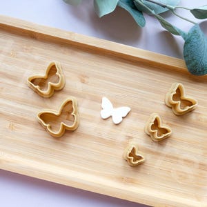 Puede incluir: Una bandeja de madera contiene varios cortadores de galletas con forma de mariposa y una sola mariposa blanca. Los cortadores de galletas son de color marrón claro y la bandeja tiene una veta de madera natural. Una ramita de vegetación está en la esquina superior derecha.