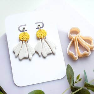 May include: A pair of handmade white and yellow daisy earrings with silver-colored hardware. The earrings are displayed on a white card, next to a daisy-shaped clay cutter. The earrings have a textured yellow circle and white petal-shaped dangles.