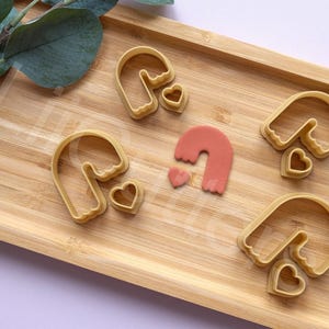 Puede incluir: Cinco cortadores de galletas en forma de arcos con corazones. Los cortadores de galletas están hechos de plástico marrón claro y están dispuestos sobre una superficie de madera.