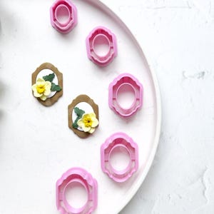 May include: A white plate with five pink plastic cookie cutters and two brown paper cutouts with yellow flowers. The cookie cutters are in the shape of a circle with a decorative edge. The paper cutouts are in the shape of a rectangle with a decorative edge.