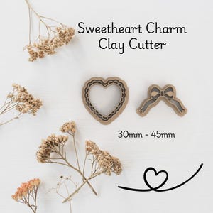 Può includere: Due taglia pasta in argilla: un cuore con bordo smerlato e un fiocco. Il testo recita "Sweetheart Charm Clay Cutter" e "30mm - 45mm". I taglia pasta sono marrone chiaro e su sfondo bianco.