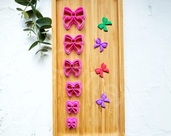 May include: A wooden board with a variety of pink, red, green, and purple bow-shaped cookie cutters. The cookie cutters are arranged in a vertical line from largest to smallest.