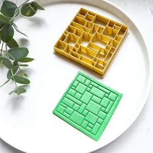 May include: Two plastic cookie cutters on a white plate. One cutter is yellow and has a geometric pattern. The other cutter is green and has a brick pattern.