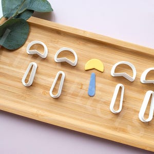 May include: A set of eight white plastic cookie cutters shaped like mushrooms and a small blue and yellow fondant cutter. The cutters are arranged on a wooden surface.
