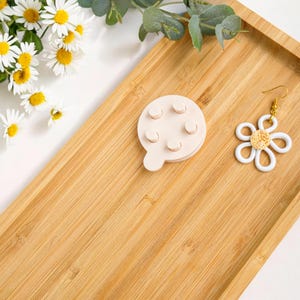 May include: A wooden tray with a white daisy-shaped earring and a small wooden object with five holes. The tray is made of light brown wood and has a natural finish.