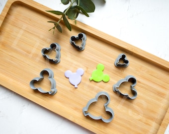May include: A set of eight grey plastic cookie cutters on a wooden surface. The cutters are shaped like Mickey Mouse heads and balloons. There are two green and two purple fondant shapes on the surface.