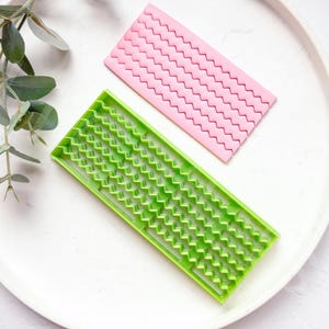 Puede incluir: Dos herramientas de decoración de pasteles de plástico, una verde y otra rosa, con un patrón ondulado. Las herramientas están en un plato blanco.