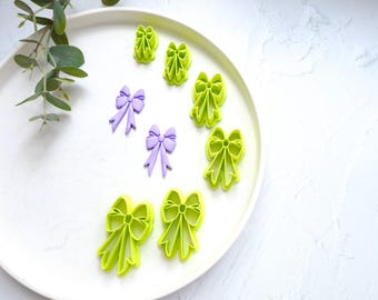 May include: A set of eight cookie cutters in the shape of bows. The cutters are made of green and purple plastic. The cutters are arranged on a white plate.