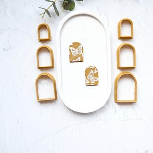 May include: A set of six gold arch-shaped cookie cutters, arranged in three pairs of different sizes, with two small yellow and white butterfly-patterned fondant pieces on a white surface.