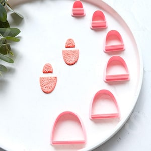 Puede incluir: Un plato blanco con pendientes de arcilla polimérica rosa y cortadores de galletas de plástico rosa en forma de arcos.