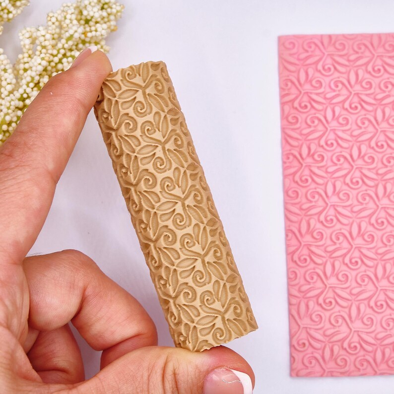 May include: A tan-colored rolling pin with a floral pattern is held in a hand, next to a pink, patterned sheet. The rolling pin has a repeating design of leaves and swirls. The pink sheet has a similar, embossed floral pattern.