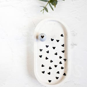 May include: White oval tray with black Mickey Mouse shapes scattered across the surface. A small gray ceramic Mickey Mouse head sits in the center of the tray.