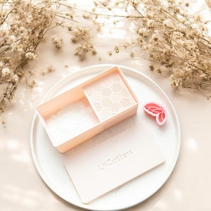 Puede incluir: Un recipiente rectangular rosa claro con un inserto con estampado geométrico, un cortador en forma de mariposa rosa y una herramienta rectangular rosa claro con el texto "LilCutters" en un plato blanco. Flores secas en el fondo.