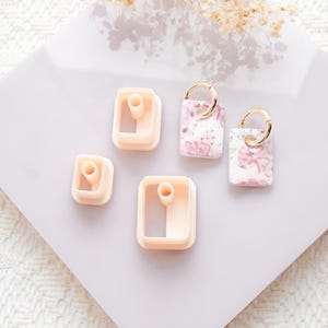 Puede incluir: Una colecci&oacute;n de cortadores y pendientes de arcilla polim&eacute;rica. Los cortadores tienen forma cuadrada y rectangular, en color melocot&oacute;n claro. Los pendientes tienen un estampado floral en rosa y blanco, con aros dorados. Los art&iacute;culos est&aacute;n dispuestos sobre una superficie lila claro.