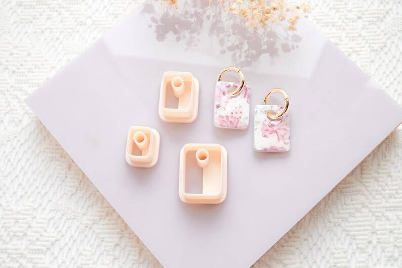 Puede incluir: Una colecci&oacute;n de cortadores y pendientes de arcilla polim&eacute;rica. Los cortadores tienen forma cuadrada y rectangular, en color melocot&oacute;n claro. Los pendientes tienen un estampado floral en rosa y blanco, con aros dorados. Los art&iacute;culos est&aacute;n dispuestos sobre una superficie lila claro.