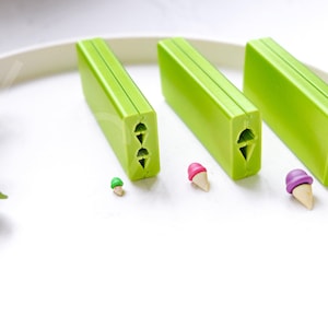 Könnte beinhalten: Drei grüne Kunststoffformen zum Herstellen von Eiswaffeln in verschiedenen Größen. Die Formen haben eine Reihe von drei verschiedenen Kegelformen. Es gibt drei kleine Eiswaffeln, die mit den Formen hergestellt wurden, eine grüne, eine rosa und eine violette.