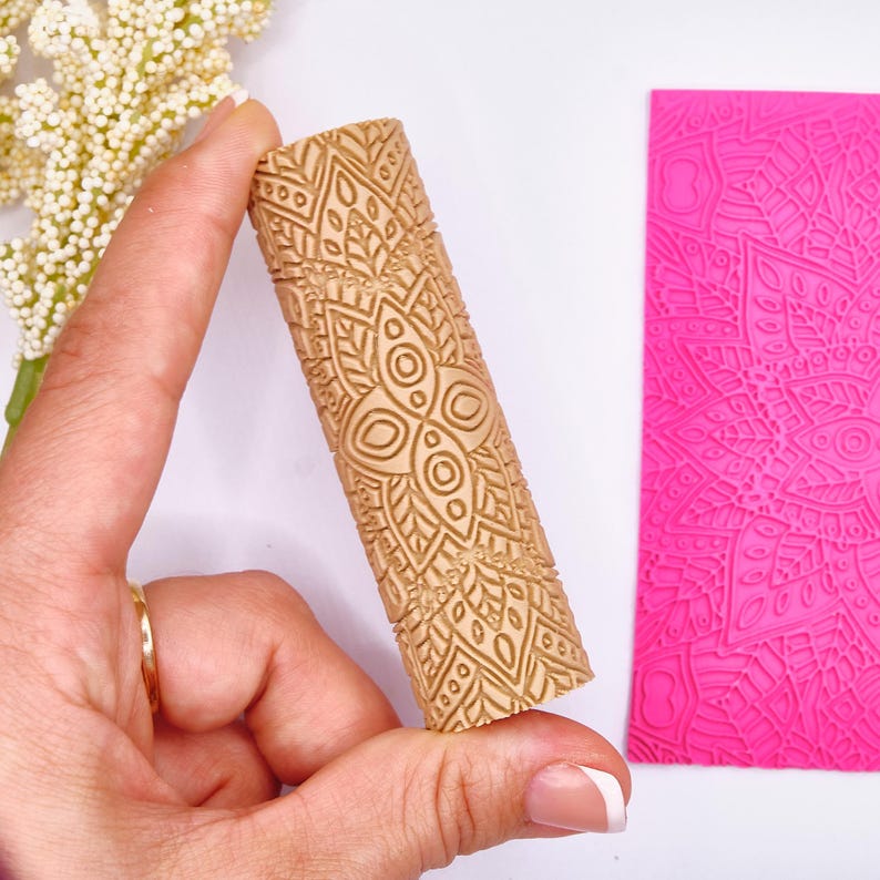 May include: A light brown, cylindrical rolling pin with a detailed, symmetrical floral pattern. The rolling pin is held next to a bright pink, rectangular sheet with a matching embossed design. The design features circles, leaves, and geometric shapes.