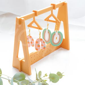 Può includere: Tre paia di orecchini fatti a mano esposti su un supporto in legno giallo. Gli orecchini sono realizzati in pasta polimerica e presentano diversi design, tra cui un motivo pesca e arancia, un design a strisce bianche e verdi e un design a strisce multicolori.