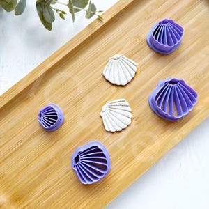 Op de afbeelding: Vijf paarse plastic koekjesvormen in de vorm van schelpen op een houten oppervlak. De vormen zijn van verschillende groottes en hebben een gedetailleerd ontwerp.