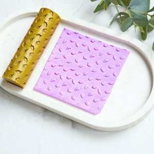 May include: A gold rolling pin with a leaf pattern and a purple silicone sheet with a matching leaf pattern. The rolling pin and sheet are on a white oval tray.