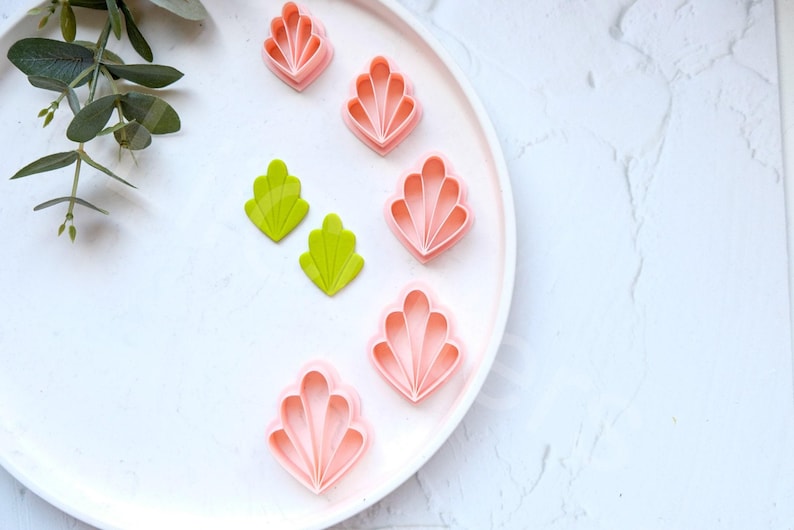 May include: A white plate with five pink and two green plastic cookie cutters shaped like seashells and leaves.