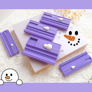 Peut inclure: Plusieurs moules à savon violets avec des épingles décoratives, avec des accents blancs en forme de bonhomme de neige. L'image comprend une illustration de visage de bonhomme de neige et un dessin animé de bonhomme de neige. Les moules sont disposés sur une surface blanche.