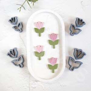 May include: A set of five cookie cutters in the shape of flowers. The cutters are made of a light gray plastic and are arranged on a white surface. The flowers have pink petals and green stems.